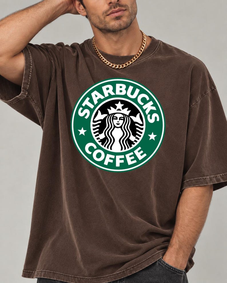 May include: A brown short-sleeve t-shirt featuring a large, circular green Starbucks Coffee logo printed on the chest. The logo includes the iconic siren graphic, white text, and two white stars on a green background. The shirt has a crew neckline.