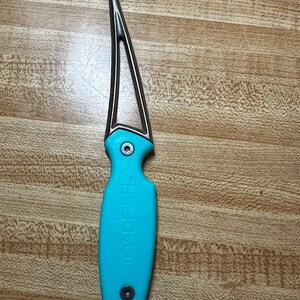 May include: A seafood tool with a curved, pointed metal tip and a bright turquoise handle. The handle features the embossed text TOADFISH and is secured with two silver-toned rivets. The tool has an open, triangular design near the base of the metal.