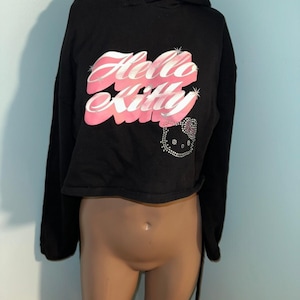 May include: A black cropped hoodie featuring the text Hello Kitty in a stylized pink and white font across the chest. Below the text is a small graphic of the Hello Kitty character face outlined in silver rhinestones with a matching bow detail.