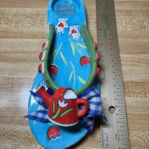Könnte beinhalten: Eine blaue, flip-flop-förmige Keramikschale mit einer Länge von etwa 24 cm. Sie hat grüne Riemen, ein rotes Gießkannen-Ornament, eine blau-weiß karierte Schleife und den Schriftzug 'diane's Happy Toes' im Fersenbereich.