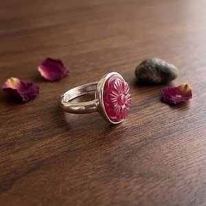 May include: A silver ring featuring a polished, deep pink gemstone carved into a floral shape. The ring has a smooth, metallic band and is set with a circular, textured stone that displays intricate petal-like engravings on its surface.