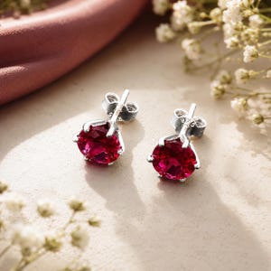 Puede incluir: Un par de pendientes de botón con piedras redondas facetadas de color rosa intenso engastadas en garras de metal plateado. Cada pendiente tiene un cierre de presión y lleva el sello 925, lo que indica que están hechos de plata de ley.
