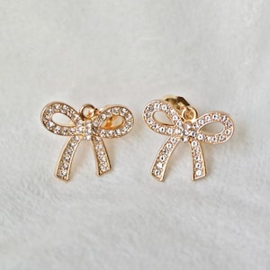 May include: A pair of gold-colored bow-shaped stud earrings. Each earring is encrusted with small, clear crystals that outline the bow shape. The earrings are displayed on a soft, white surface.