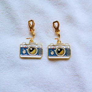 May include: A pair of gold camera-shaped earrings with blue and white enamel details. The earrings have a clip-on closure.