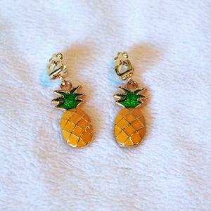 May include: A pair of gold-tone clip-on earrings featuring a pineapple design. The pineapples are yellow with green tops and are outlined in gold.