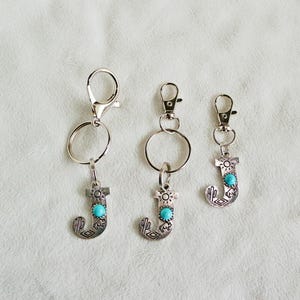 May include: Three silver keychains with the letter "J" pendants. Each pendant has a turquoise-colored stone and a decorative design. The keychains are of varying sizes and have a lobster clasp or a split ring.