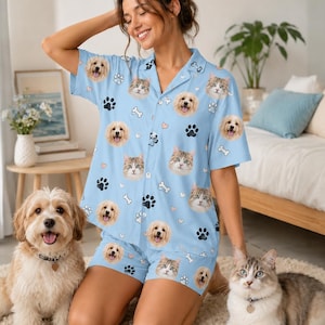 May include: A light blue pajama set consisting of a short-sleeved button-down shirt and shorts, featuring an all-over print of dog and cat faces, black paw prints, white bones, and small pink hearts on a soft fabric.