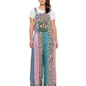 May include: A woman wearing a white t-shirt under a colorful jumpsuit. The jumpsuit features a patchwork design with floral and geometric patterns in shades of blue, pink, green, and cream. The wide-leg pants have a decorative border at the hem.