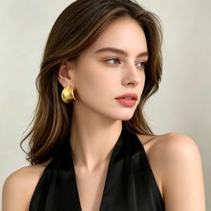 May include: A gold-toned earring with a smooth, curved, semi-circular shape. The earring features a polished metallic finish that reflects light, creating a subtle sheen across its surface. It is designed to sit flush against the earlobe.