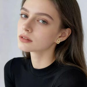 May include: A gold stud earring featuring an oval shape with a horizontal bar across the center. The earring has a textured, beaded surface finish and is worn on the earlobe. The design is metallic gold in color and has a small, compact size.