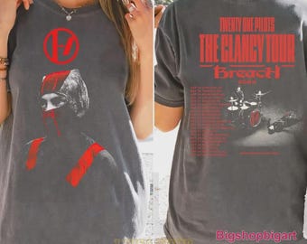 May include: A charcoal gray t-shirt featuring a red graphic of a masked figure on the front with a circular logo. The back displays red text reading Twenty One Pilots The Clancy Tour Breach 2025 above a monochromatic image of a drum kit.