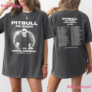 May include: A charcoal gray short-sleeve t-shirt featuring a graphic of Pitbull with the text "PITBULL I'M BACK NORTH AMERICA LIL JON". The back of the shirt displays a list of tour dates and locations for the North America tour with white text.
