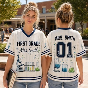 May include: A cream-colored V-neck jersey with navy blue pinstripes and trim. The front features the text FIRST GRADE Mrs. Smith, a microscope, test tubes, and a flask. The back displays MRS. SMITH, the number 01, and science equipment graphics.