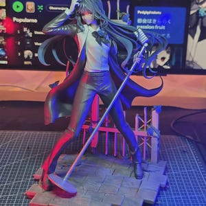 Op de afbeelding: Een gedetailleerd anime-figuur met lang donker haar, gekleed in een wit overhemd, zwarte stropdas en een lange zwarte jas. Het figuur staat op een voetstuk met steentextuur en een hek, en houdt een zilveren microfoonstandaard vast.