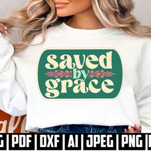 May include: A white crewneck sweatshirt featuring a rectangular green graphic with rounded corners. The text "saved by grace" is printed in cream-coloured letters, with a small pink floral design separating the words "saved" and "grace".