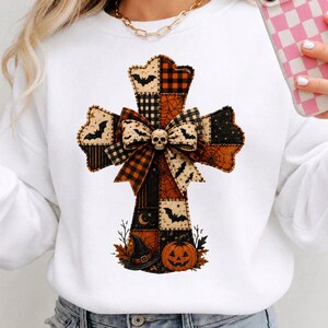May include: A white long-sleeve sweatshirt featuring a large, central cross graphic composed of patchwork patterns in orange, black, and cream. The cross includes bat silhouettes, spider webs, a central bow, a skull, a witch hat, and a pumpkin.