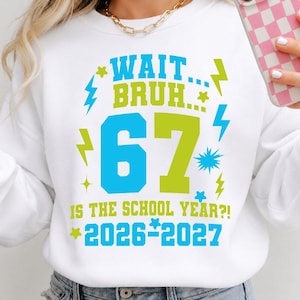 Op de afbeelding: Een wit sweatshirt met ronde hals en een grafische print in blauw en limoengroen. De tekst luidt "WAIT... BRUH... 67 IS THE SCHOOL YEAR?! 2026-2027", omringd door bliksemschicht- en sterpictogrammen. Het ontwerp staat centraal op de voorzijde.