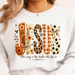 Op de afbeelding: Een wit sweatshirt met ronde hals en het woord JESUS in grote, gestileerde letters met diverse patronen zoals oranje bloemen, luipaardprint, oranje ruitjes en zwarte stippen, met daaronder de tekst the way the truth the life JOHN 14:6.