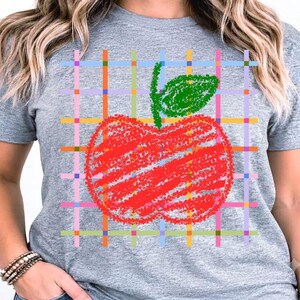 Peut inclure: Un t-shirt gris chiné à manches courtes avec un motif central représentant une pomme rouge surmontée d'une feuille verte. La pomme est superposée à un quadrillage coloré composé de fines lignes bleues, jaunes, orange et roses.