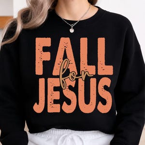Könnte beinhalten: Ein schwarzes Sweatshirt mit Rundhalsausschnitt und dem Text FALL FOR JESUS in einer verwaschenen, pfirsichfarbenen Blockschrift. Das Wort FOR ist in einer kleineren, golden-braunen Schreibschrift über die Mitte des Textes gelegt.