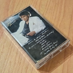 May include: A clear plastic cassette tape case featuring the Michael Jackson Thriller album cover. The artwork shows Michael Jackson in a white blazer and black shirt. The tracklist is printed in white cursive text on a black background.