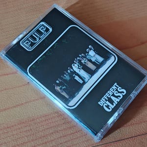 May include: A clear plastic cassette tape case featuring the album Different Class by the band Pulp. The cover art is black with white text and a central rectangular image showing a group of people in formal attire standing in a line.