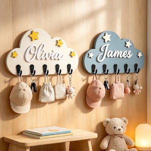 May include: Two cloud-shaped wall hooks made of wood, one cream with yellow stars and the name Olivia, the other blue with white stars and the name James. Each features five black metal hooks holding items like hats, small bags, and keychains.