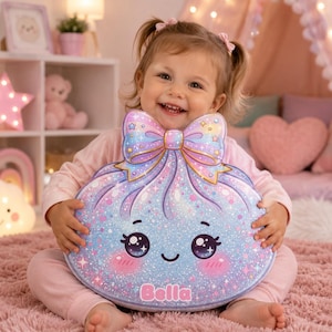 May include: A plush cushion shaped like a rounded pouch with a large bow on top. The cushion features a light blue and purple gradient with a glittery finish, a smiling face with large dark eyes, pink cheeks, and the name Bella printed in pink.