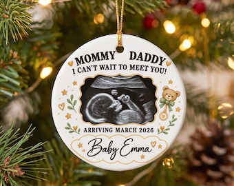 Può includere: Un ornamento rotondo in ceramica bianca con un'ecografia in bianco e nero al centro. Il testo recita: MOMMY DADDY I CAN'T WAIT TO MEET YOU! ARRIVING MARCH 2026 e Baby Emma. Decorato con stelle dorate, foglie verdi e un piccolo orsacchiotto.