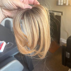 May include: A hair topper featuring multi-tonal blonde strands with darker roots. The hairpiece has a smooth, layered texture with a natural gradient from a deep brown base at the crown transitioning into lighter golden and platinum blonde highlights.