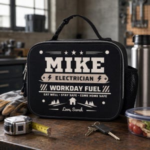 May include: A black rectangular lunch bag with a top handle and side mesh pocket. The front features white text reading MIKE ELECTRICIAN WORKDAY FUEL EAT WELL STAY SAFE COME HOME SAFE Love, Sarah, along with graphics of stars, lightning bolts, and a house.