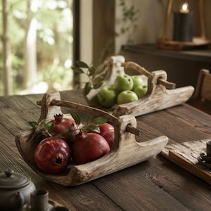 May include: Two rustic, elongated wooden bowls with arched handles, each carved from light-toned wood. The front bowl holds three deep red pomegranates with small green leaves, while the back bowl contains several bright green apples.