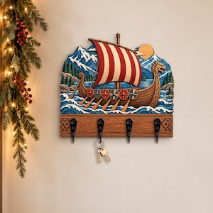 Op de afbeelding: Een houten wand-sleutelhouder met een uitgesneden Vikingschip met een rood-wit gestreept zeil, blauwe golven en besneeuwde bergen. De basis heeft vier zwarte metalen haken, waaraan een zilveren sleutelbos aan de tweede haak hangt.