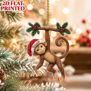 May include: A 2D flat printed Christmas tree ornament featuring a brown monkey hanging from a branch with green holly leaves and red berries. The monkey wears a red Santa hat with the name Thomas printed on it, suspended by a gold string.