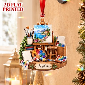 Può includere: Un ornamento sospeso stampato in 2D piatto che raffigura una scena di studio d'artista con cavalletto, dipinto di paesaggio costiero, pennelli, tavolozza, sgabello e una targhetta con il nome Sophia. Il design presenta colori vivaci e articoli d'arte.