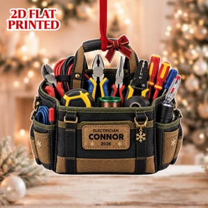 May include: A 2D flat printed hanging ornament shaped like a black and brown tool bag filled with various tools including pliers, screwdrivers, and a tape measure. A label on the front reads ELECTRICIAN CONNOR 2026 with snowflake icons.