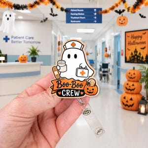 May include: A badge reel featuring a white ghost wearing a nurse cap, holding a roll of gauze and a medical kit. The design includes a bandage, a pumpkin, and the text Boo-Boo CREW in black lettering on an orange banner with a glittery finish.