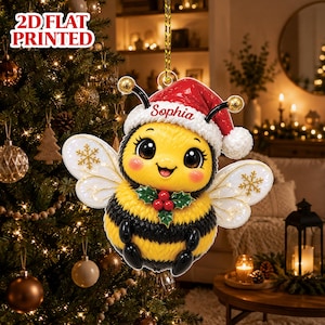 May include: A 2D flat printed Christmas tree ornament featuring a yellow and black bee wearing a red Santa hat with the name Sophia written on the white brim. The bee has large eyes, white wings with gold snowflake accents, and a holly berry sprig.