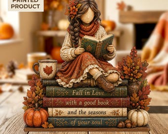 May include: A 2D flat printed decorative figurine featuring a person with long brown hair adorned with autumn leaves, wearing a cream sweater and orange scarf, reading a green book while sitting on a stack of four books with the text Fall in Love with a good book and the seasons of your soul, accented by pumpkins and fall foliage.