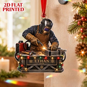 Puede incluir: Un adorno navideño plano en 2D que muestra a un soldador con casco protector y delantal marrón trabajando en un banco de trabajo mientras saltan chispas. El banco está decorado con luces de colores y muestra el texto ETHAN 2026.