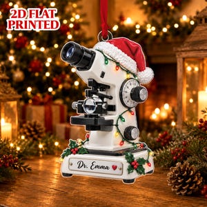 Peut inclure: Une décoration plate 2D en forme de microscope blanc portant un bonnet de Père Noël rouge et blanc. Le microscope est entouré d'une guirlande lumineuse colorée et décoré de feuilles de houx. Une plaque porte l'inscription Dr. Emma avec un cœur.