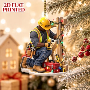 Può includere: Un ornamento sospeso stampato in 2D piatto raffigurante un operaio edile con casco giallo con la scritta 2026 ed ETHAN, gilet ad alta visibilità, jeans blu e stivali da lavoro, che usa un trapano su un palo di legno decorato con luci natalizie.