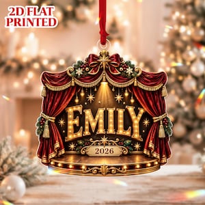 May include: A flat, 2D printed Christmas ornament featuring a theater stage design with deep red curtains, gold tassels, and a wooden floor. The name EMILY is displayed in large, illuminated marquee-style letters above the year 2026.