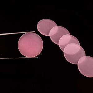 May include: A set of six translucent, circular, light pink discs arranged in a curved line. One disc is held by a pair of thin, silver-colored metal tweezers, while the other five discs are positioned behind it against a solid black background.