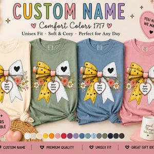 Könnte beinhalten: Vier T-Shirts in Creme, Salbeigrün, Blau und Rosa, jeweils mit einer Grafik einer gelben Schleife im Bleistiftmuster und einem herzförmigen Anhänger mit der Aufschrift 'your name here', umgeben von kleinen Blumen und Herzen, mit Text 'CUSTOM NAME'.