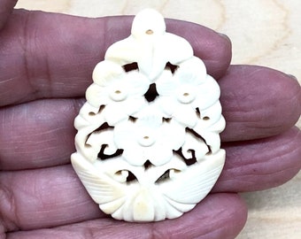 May include: A white carved bone pendant with a floral design. The pendant has a hole at the top for stringing.