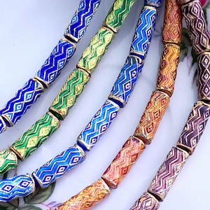 Può includere: Primo piano di una collana di perline di vetro colorate con motivi geometrici. Le perline sono cilindriche e hanno un bordo dorato. Le perline sono disposte a forma di arcobaleno, con perline blu, verdi, arancioni e viola.