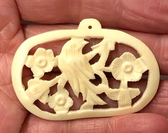 May include: A carved ivory pendant with a floral design. The pendant is oval shaped and has a hole at the top for hanging.