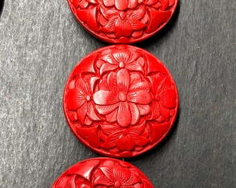 Può includere: Primo piano di diversi oggetti decorativi rotondi di colore rosso brillante. Ogni oggetto presenta un design floreale dettagliato intagliato sulla sua superficie. Gli oggetti sono disposti in fila su uno sfondo scuro, evidenziando i loro motivi.