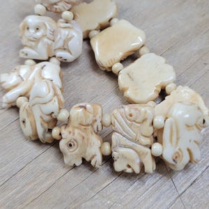 May include: Ivory-colored bracelet featuring carved animal figures and irregular-shaped beads. The bracelet includes a bird, monkey, pig, buffalo, and other animals. The bracelet is strung together with small, round beads.